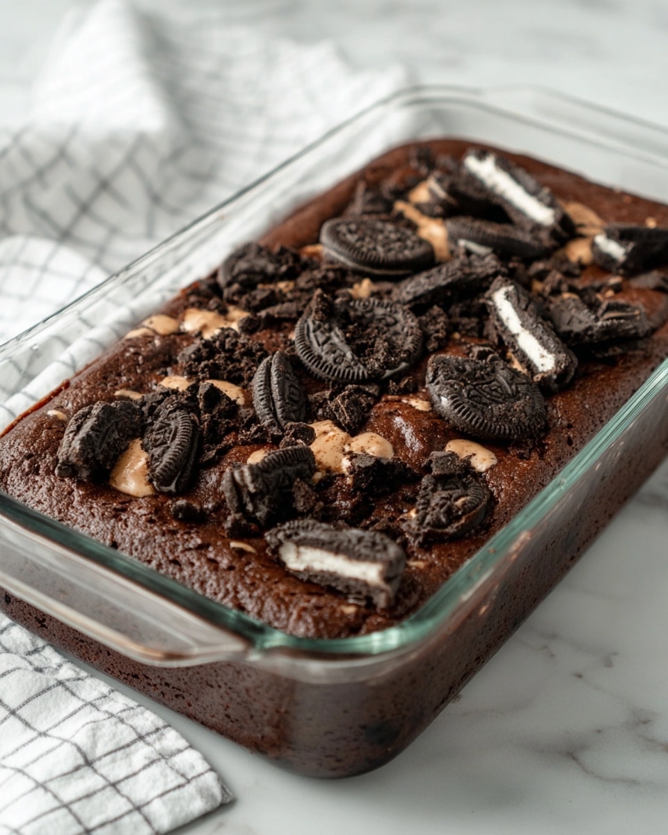 The image shows a rectangular glass baking dish filled with a thick, dark brown chocolate cake that has a rich and slightly cracked surface. The top layer is decorated with chunks of black and white sandwich cookies, some whole and some broken, scattered unevenly across the cake. The cookies add texture and contrast to the smooth chocolate base. The dish is placed on a white marbled surface, and a white kitchen towel with a light grid pattern is visible in the background. photo taken with an iphone --ar 4:5 --v 7