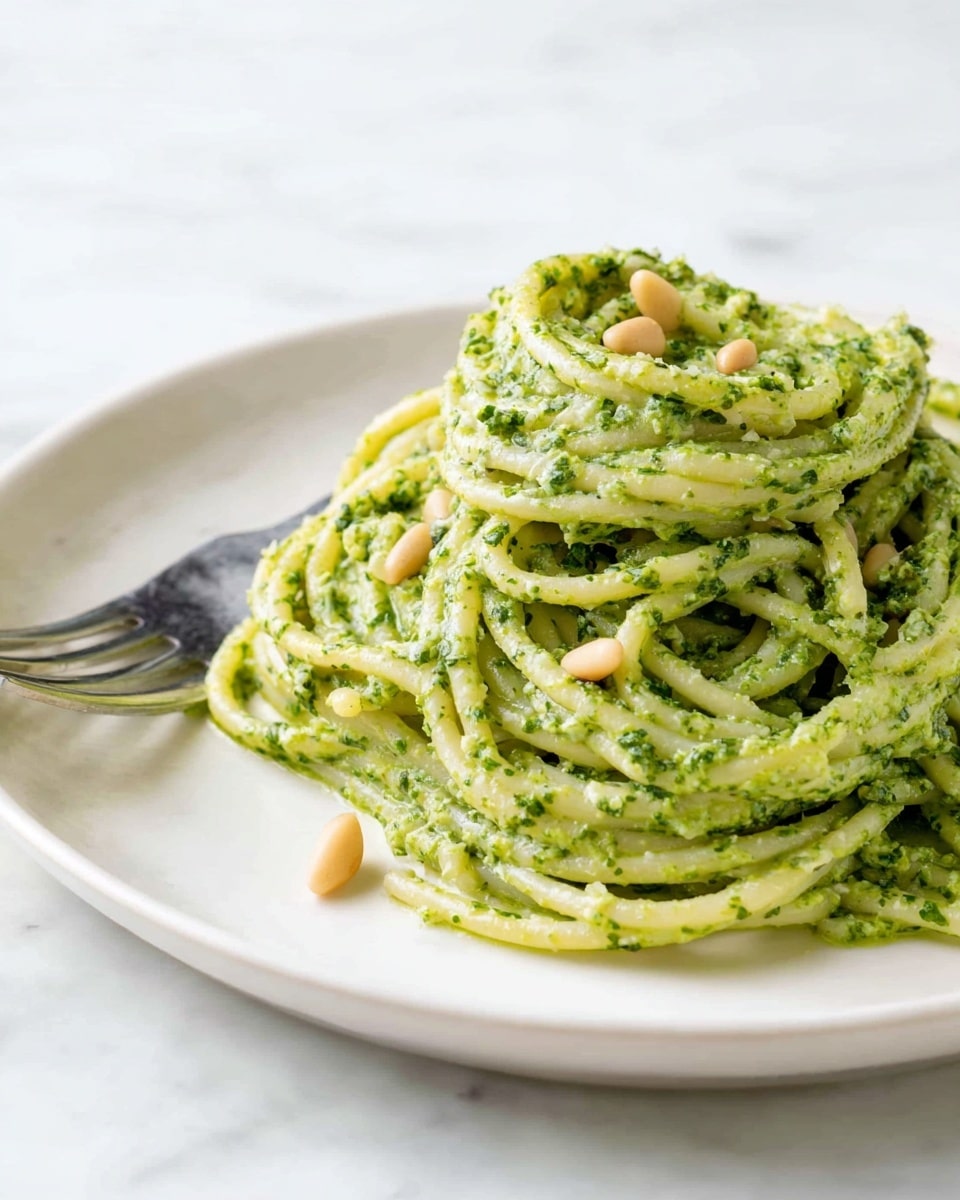 A white plate holds a twisted pile of spaghetti coated in a creamy green pesto sauce with visible bits of herbs. Scattered pine nuts add small touches of beige on top. A silver fork is partly visible behind the pasta, with its prongs resting on the plate. The background shows a soft white marbled texture that contrasts the vibrant green pasta. photo taken with an iphone --ar 4:5 --v 7