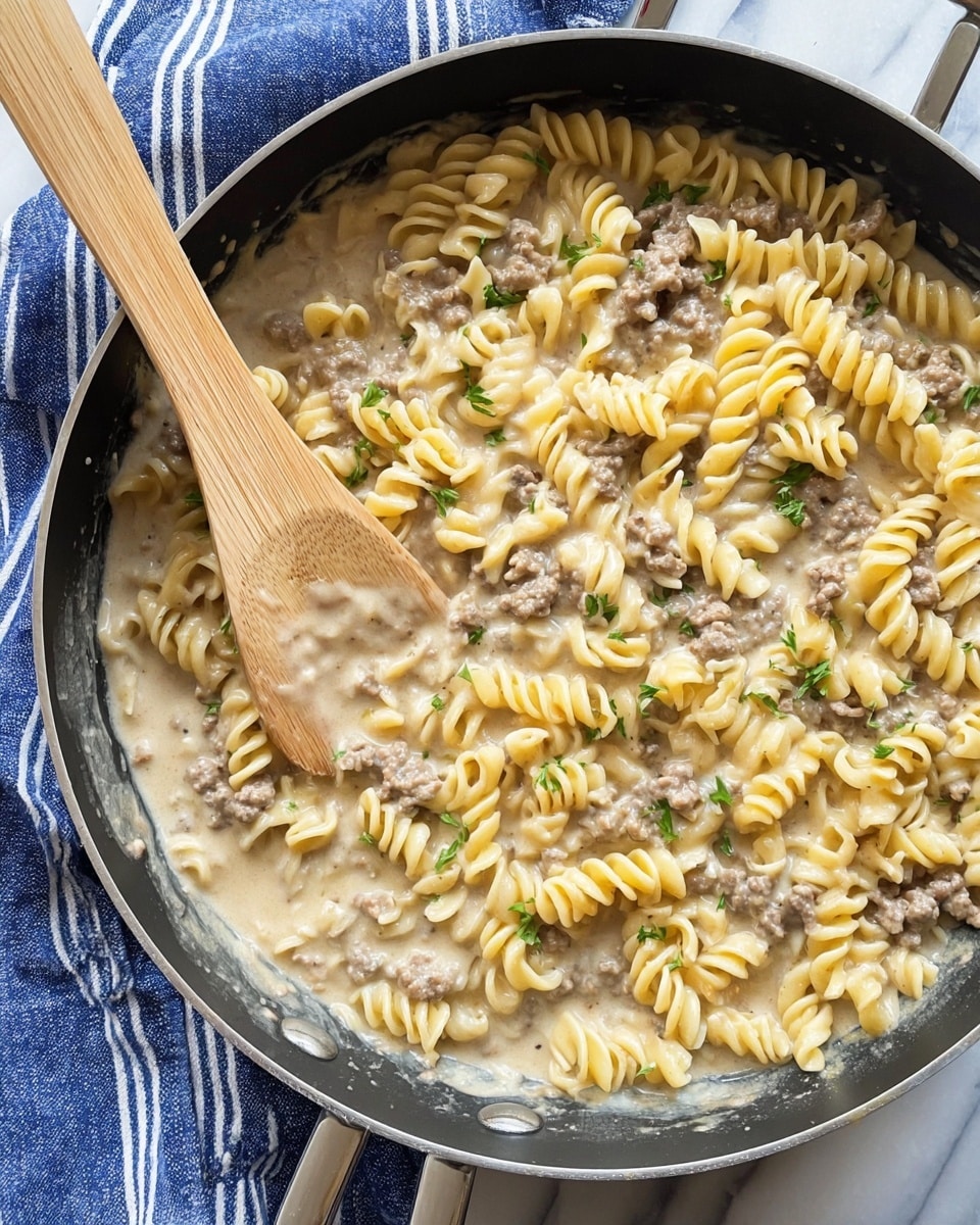 A close-up view of a black pan filled with a creamy pasta dish showing about three layers: a base layer of small spiral yellow pasta, a middle layer of light brown ground meat mixed in, and a top layer of creamy light beige sauce coating the pasta and meat evenly, with small pieces of onion and green parsley scattered on top. A wooden spatula rests inside the pan, partially stirring the pasta. The pan is on a white marbled surface with a blue and white striped cloth partly visible on the side. Photo taken with an iphone --ar 4:5 --v 7
