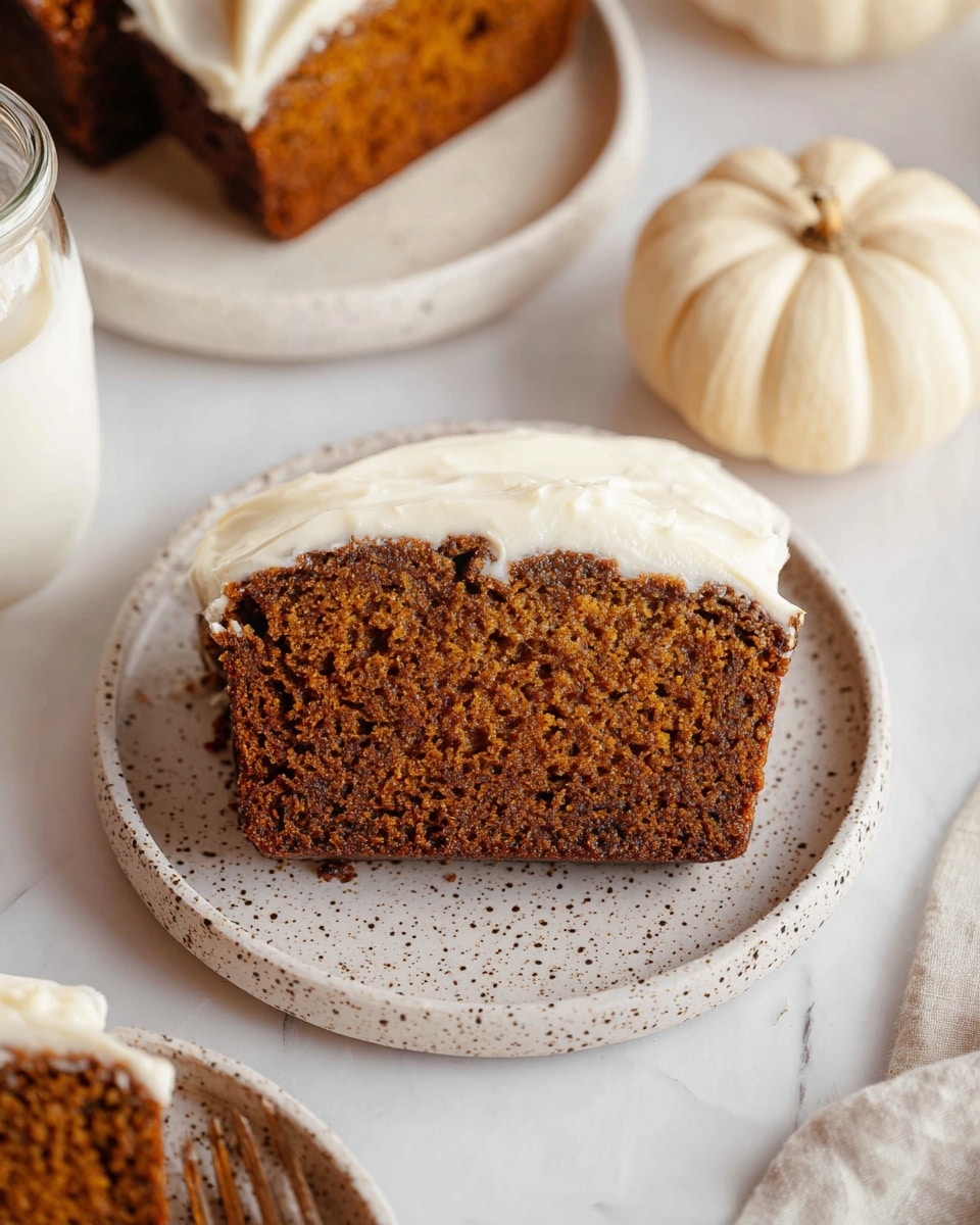 A single slice of moist, dark brown pumpkin bread with a slightly rough crumb texture sits on a round white plate with speckled black dots. The bread is topped with a thick layer of smooth, creamy white frosting that covers one side of the slice. The scene includes the edge of another similar plate with a slice of the same bread and frosting in the background, a small white pumpkin, and a glass jar containing white milk, all placed on a white marbled surface. Photo taken with an iphone --ar 4:5 --v 7