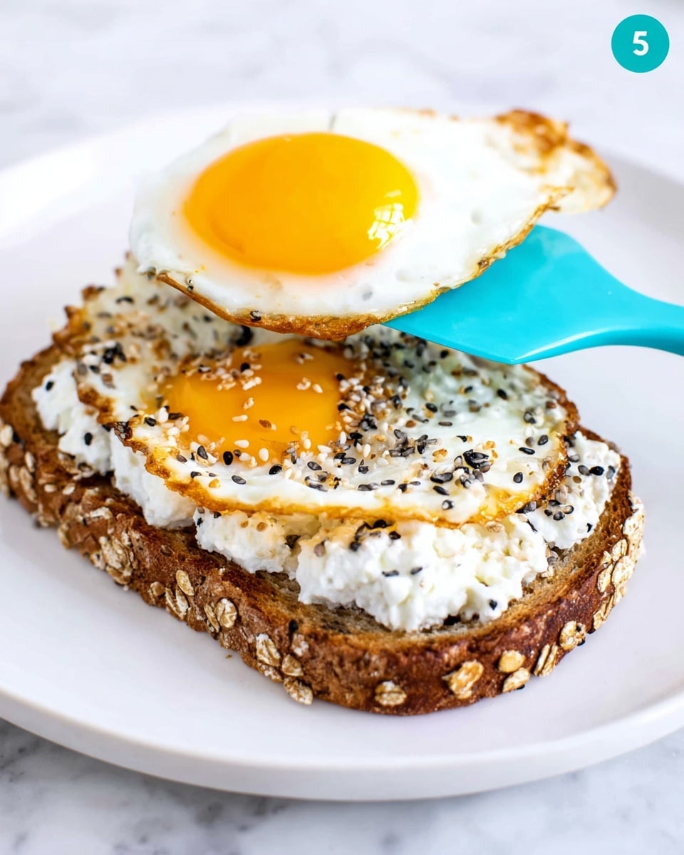 A close-up image of a single slice of toasted brown whole grain bread with visible oats on the crust, topped with a thick, creamy layer of white cottage cheese sprinkled with a mix of black and white seeds. Above the toast, a blue spatula holds a sunny-side-up egg with a bright yellow yolk and edges of crispy, golden brown cooked egg white. The plate underneath is white, resting on a white marbled surface. photo taken with an iphone --ar 4:5 --v 7
