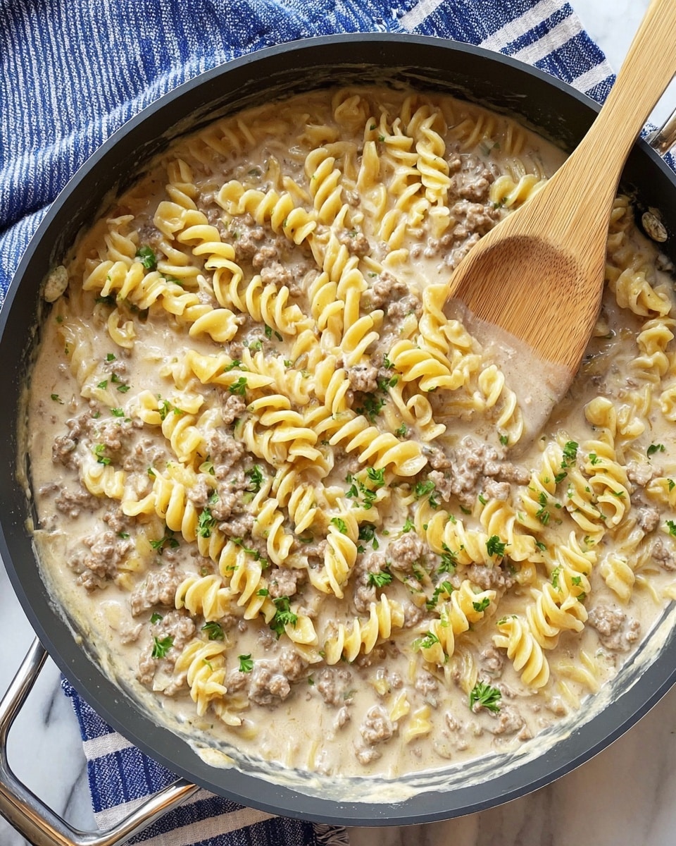 This image shows a pan filled with creamy pasta. The bottom layer is a light beige sauce with a smooth, thick texture, covering the pan evenly. On top of the sauce, there are medium-sized spiraled pasta pieces in a pale yellow color mixed with small bits of cooked ground meat in brown. Small green parsley leaves are scattered across the pasta, adding a fresh touch. A light wooden spatula with a smooth surface is placed in the pan, partially submerged in the pasta, stirring the mixture. The pan has black sides with silver handles. The setting shows a white marbled surface underneath with a blue and white striped cloth beside the pan. Photo taken with an iphone --ar 4:5 --v 7