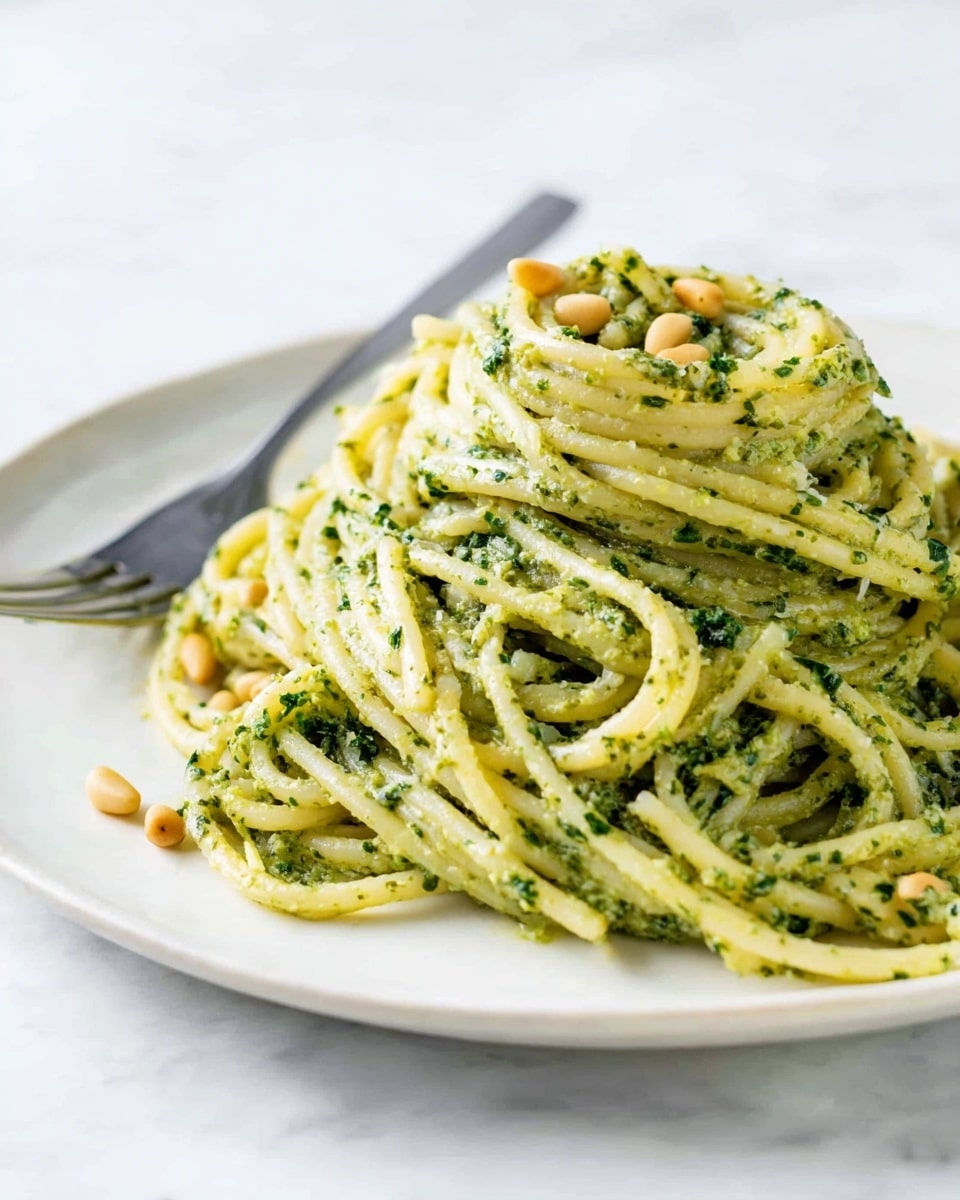 A round white plate with a small pile of spaghetti pasta coated in a creamy green pesto sauce, the sauce visibly mixed with tiny bits of herbs. The pasta is twisted and layered in a loose spiral shape around itself, creating depth and texture. Scattered lightly on top are small beige pine nuts adding slight contrast. A silver fork is placed on the plate behind the pasta, resting against the edge. The backdrop is a smooth white marbled surface that gives a clean and bright look. Photo taken with an iphone --ar 4:5 --v 7