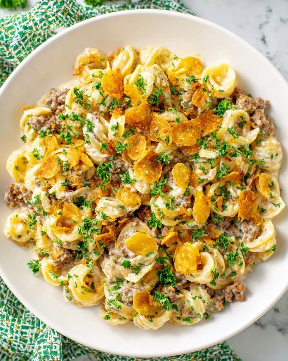 A white round plate filled with one layer of creamy pasta, mixed with small pieces of browned ground meat and crispy golden potato chips scattered evenly throughout. The pasta is pale yellow with a thick creamy coating, and bright green chopped parsley is sprinkled generously on top, adding a fresh color contrast. The dish sits on a white marbled surface with a green and white patterned cloth partially visible in the background. photo taken with an iphone --ar 4:5 --v 7