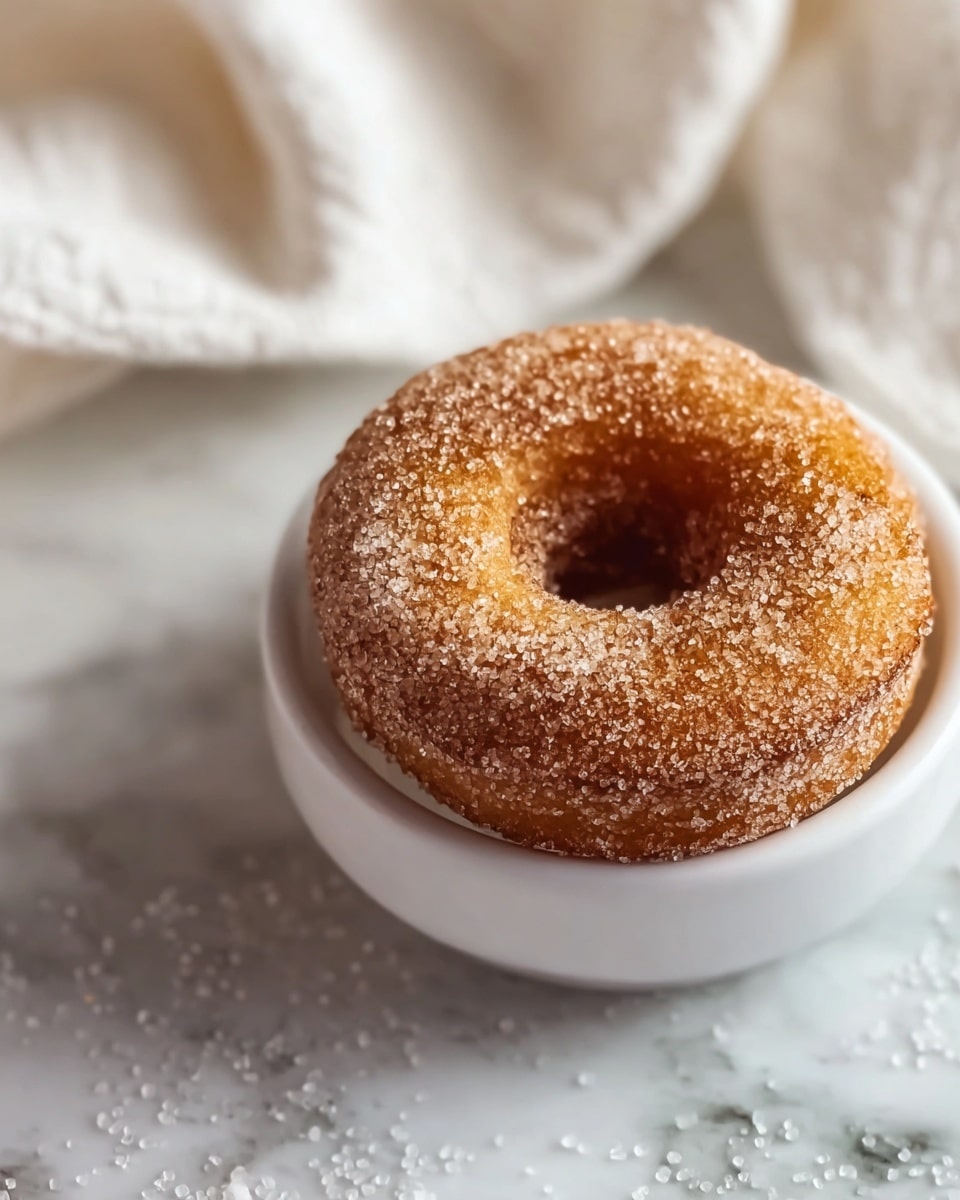 A single sugar-coated donut sits in a white bowl, with its golden brown surface covered in sparkling coarse sugar crystals. The donut's texture looks soft and slightly crispy on the edges. The bowl rests on a white marbled surface scattered with sugar grains, and a blurred white towel is in the background. The light highlights the sugar crystals, making them glisten. photo taken with an iphone --ar 4:5 --v 7