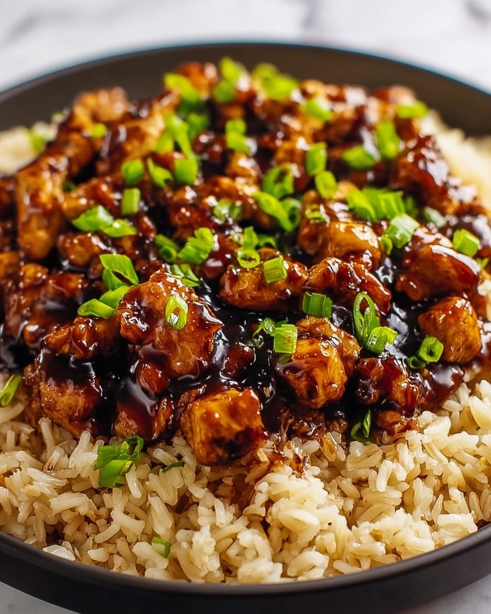 The dish shows two main layers: the bottom layer is light brown cooked rice with a slightly oily shininess and some grains sticking together, filling the whole round white plate. On top, there is a thick layer of small, glossy chicken pieces, dark brown with a rich shiny sauce coating them, giving a caramelized look. Scattered on the chicken, there are small, bright green chopped spring onions that add a fresh touch. The food is placed on a white marbled surface. photo taken with an iphone --ar 4:5 --v 7