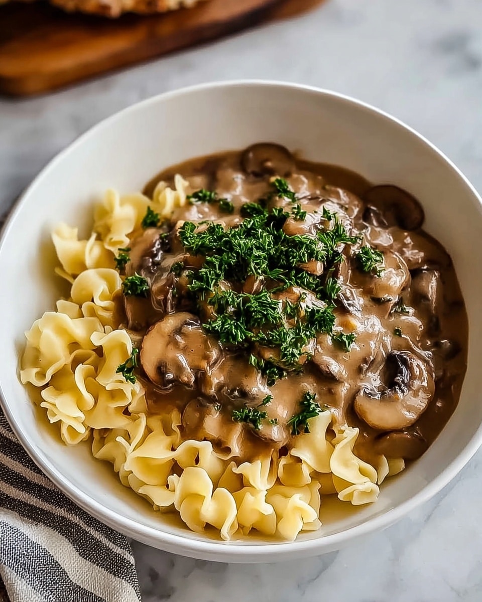 The image shows a white bowl filled with creamy macaroni and cheese on one side, which is smooth and pale yellow with small flecks of black pepper. On the other side of the bowl, there is a layer of sautéed mushrooms with a mix of dark brown and light tan colors, covered in a glossy sauce. The macaroni and cheese looks soft and cheesy, while the mushrooms add a moist, juicy texture. A few small sprigs of chopped green herbs are placed in the middle where the two layers meet. The bowl sits on a white marbled surface next to a light-colored cloth napkin and pieces of beige toasted bread. Photo taken with an iphone --ar 4:5 --v 7