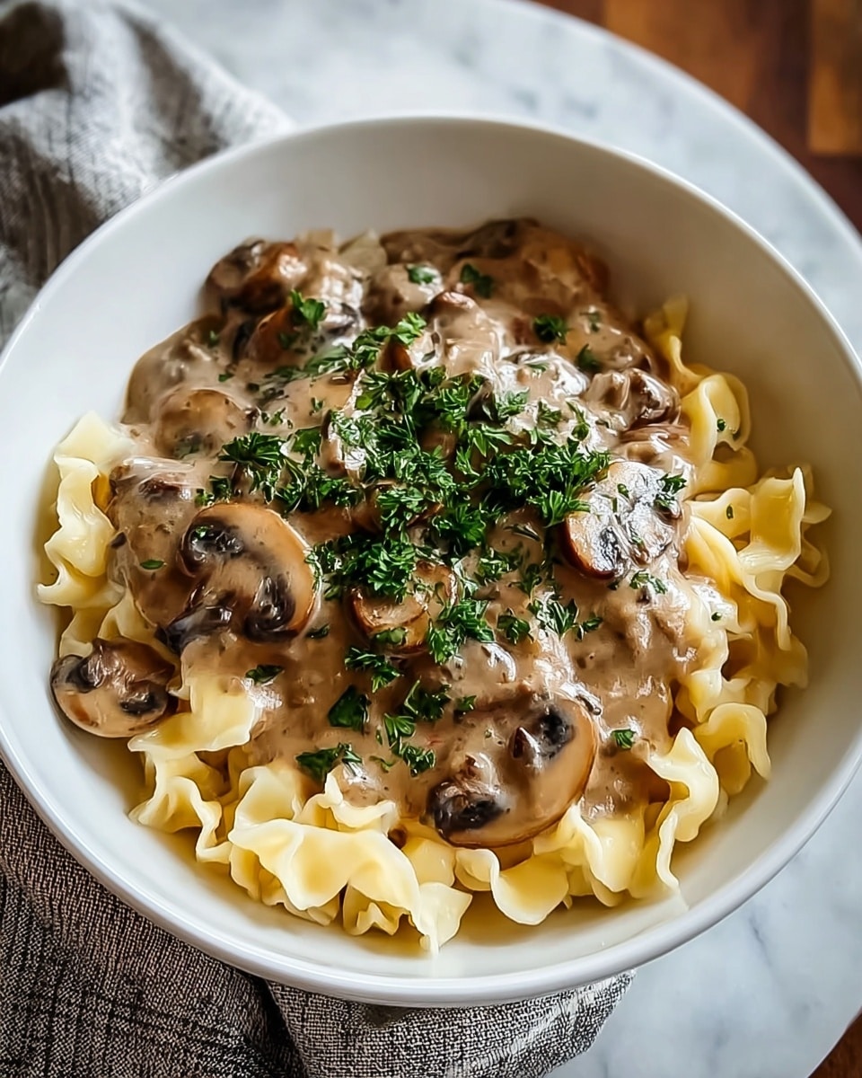 A white bowl holding three layers: at the bottom are soft, slightly folded light yellow pasta pieces; above this is a rich, creamy brown mushroom sauce with visible sliced mushrooms coated in it; the top layer is fresh finely chopped green parsley sprinkled over the sauce. The bowl sits on a white marbled surface with a cloth napkin nearby. photo taken with an iphone --ar 4:5 --v 7
