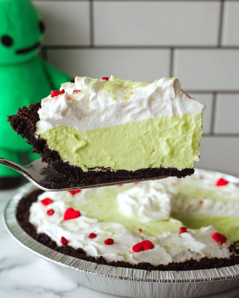 A slice of pie is held up showing three clear layers: a dark crumbly crust at the bottom, a thick bright green creamy middle layer, and a white whipped topping on top sprinkled with green powder and small scattered red heart-shaped decorations. The slice is taken from a pie in a silver foil pan, sitting on a white marbled surface, with the background of white tiled wall. A green stuffed toy is partially visible on the left side. Photo taken with an iphone --ar 4:5 --v 7