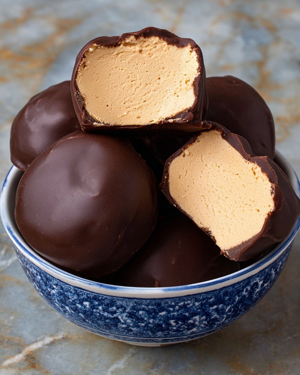 The image shows several round chocolate-covered treats placed inside a white bowl with blue patterns, sitting on a white marbled surface. There are six pieces visible, three of them whole with smooth, shiny dark brown chocolate coatings, and three cut in half, revealing a thick, light tan, creamy filling inside that looks soft and airy. The cut pieces are stacked on top of some whole pieces, showing the contrast between the outer chocolate layer and the inner beige filling. Photo taken with an iphone --ar 4:5 --v 7