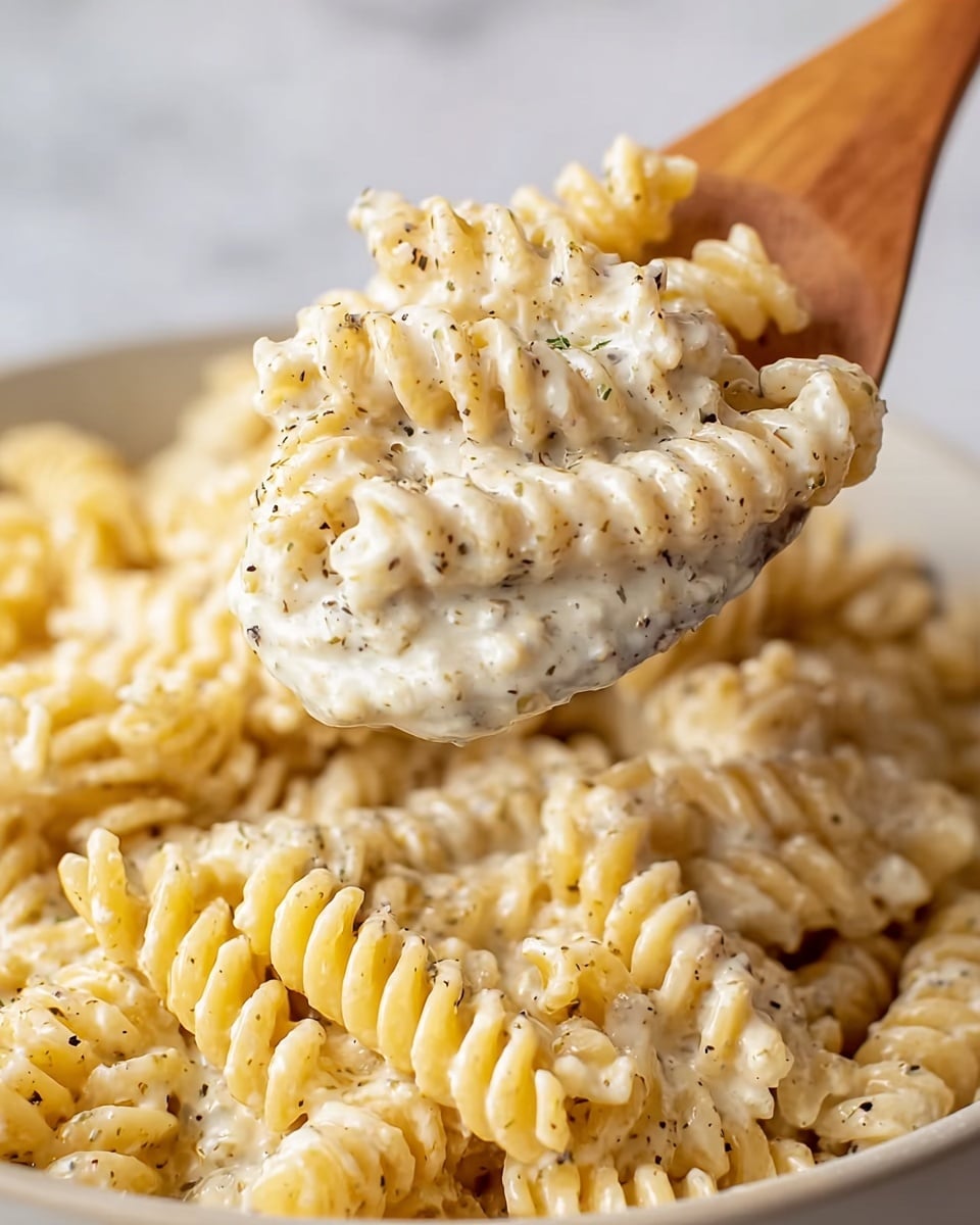 A close-up view of creamy rotini pasta covered in a thick white sauce with visible small black pepper and herb specks, showing the sauce clinging to the spiraled shapes. The creamy sauce is smooth and rich, coating the light yellow pasta evenly. A wooden spoon lifts a portion of the pasta, capturing the texture and details of both the pasta and sauce. The pasta sits in a white bowl against a white marbled background. photo taken with an iphone --ar 4:5 --v 7