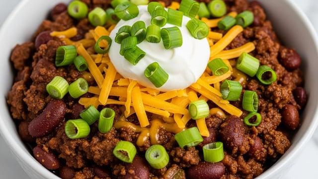 A close-up image of a bowl of chili with visible layers starting with a rich, chunky dark brown meat and kidney bean base that fills the bowl, topped with melted bright yellow shredded cheese spread evenly over the center. On top of the cheese, there is a dollop of smooth white sour cream placed near the middle, with finely chopped fresh green onions scattered generously all over the chili, cheese, and sour cream. The bowl is white with a simple design, placed on a white marbled surface. photo taken with an iphone --ar 4:5 --v 7