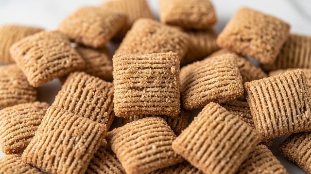 A close-up view of small, square-shaped cereal pieces coated with a crumbly mix of brown sugar and cinnamon. The cereal pieces show a rough, grainy texture with specks of cinnamon visible across their surface. The light brown color varies slightly with darker brown cinnamon spots adding depth, and the pieces are piled together, filling the frame with a soft focus on the edges and sharper detail in the middle layer. The background is a soft white marbled texture. photo taken with an iphone --ar 4:5 --v 7