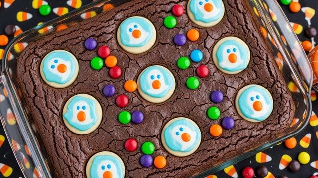 A rectangular glass baking dish holds a rich, dark, fudgy brownie layer with a cracked surface texture. Scattered evenly across the top are round sugar cookie pieces decorated with a blue and white ghost face with an orange mouth, embedded slightly into the brownie. Brightly colored candy-coated chocolates in red, green, purple, and orange are sprinkled around the cookie pieces. The dish sits on a surface covered with Halloween-themed candy and a black cloth printed with orange candy corn shapes. photo taken with an iphone --ar 4:5 --v 7