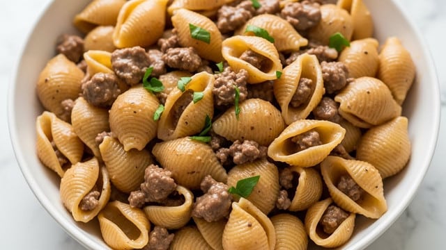 A close-up of a white bowl filled with a single layer of shell-shaped pasta coated in a light brown sauce, mixed evenly with small chunks of cooked ground beef. Scattered small green parsley leaves add a fresh touch on top, contrasting with the warm tones of the pasta and meat. The pasta shells have a ridged texture and some show their hollow centers. The scene is set against a white marbled surface. photo taken with an iphone --ar 4:5 --v 7
