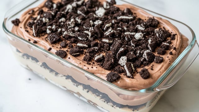 A close-up of a glass rectangular dish filled with a layered dessert. The bottom layer looks creamy and light brown, topped by a thick layer of smooth, light chocolate mousse. The mousse layer is covered generously with crushed dark chocolate cookie pieces, creating a rough, crumbly black and white texture on top. The dish is placed on a white marbled texture surface. photo taken with an iphone --ar 4:5 --v 7