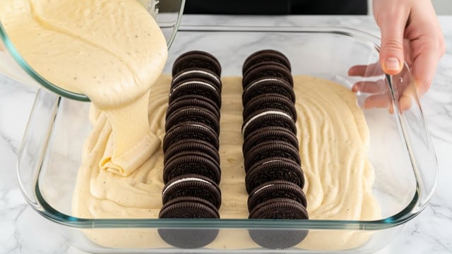 A close-up image shows two rows of Oreo cookies standing upright inside a clear rectangular glass baking dish, each sandwich cookie dark brown with white cream filling visible, positioned evenly in parallel lines; a thick, pale yellow batter with smooth texture is being poured from a glass mixing bowl by a woman's hand, coating the bottom layer and reaching between the cookies on a white marbled surface. photo taken with an iphone --ar 4:5 --v 7