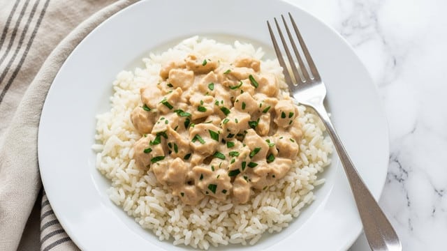 A white plate holds a bed of plain white rice as the bottom layer, topped with a creamy chicken mixture that is beige with small green herb bits evenly spread throughout. A silver fork rests on the right side of the plate. The plate is placed on a white marbled surface with a striped beige cloth partially visible on the left side. photo taken with an iphone --ar 4:5 --v 7