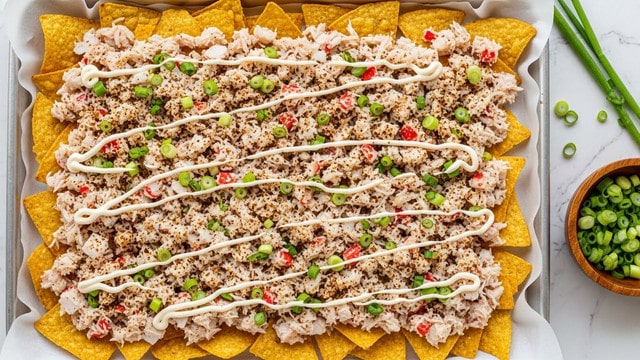 A rectangular baking tray lined with white parchment paper holds one layer of crispy golden-brown tortilla chips spread evenly at the base. On top is a thick layer of diced white and red crab meat mixed with small curls of white mayo sauce drizzled across the dish. Scattered throughout are finely chopped bright green spring onions and bits of a dark brown spice blend adding a textured look. The whole dish has colors of golden, white, red, green, and dark brown spread in an even, yet slightly messy way. The tray is placed on a white marbled surface with a wooden bowl of chopped green onions beside it. Photo taken with an iphone --ar 4:5 --v 7