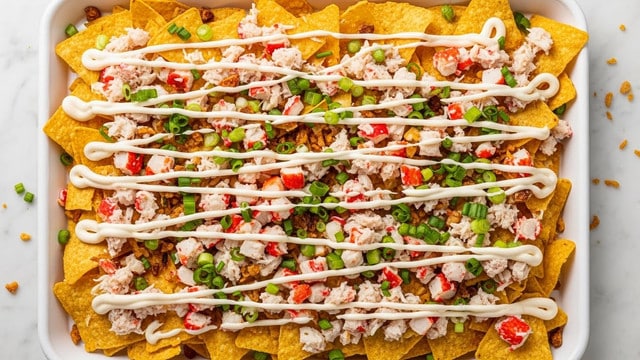 The image shows a white rectangular tray filled with multiple layers of food. The bottom layer consists of golden-brown tortilla chips spread evenly. Over the chips, there is a drizzle of white sauce applied in criss-cross lines, creating a light, smooth texture. Scattered across the surface are finely chopped pieces of red and white crab meat, adding a mix of bright red and creamy white colors. Small green chopped scallions are sprinkled evenly on top, providing fresh green pops of color. There are also small bits of crispy fried onions scattered throughout, adding dark brown crunchy texture. The tray sits on a white marbled surface with some bits of toppings fallen around the edges. The photo was taken with an iphone --ar 4:5 --v 7